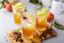 3 Gläser Apfel-Zimt-Spritz mit Eiswüfeln, Apfelscheiben. Anis und Kräutern garniert, zwei stehen auf einem Brett, daneben Äpfel und Zimtstangen.
