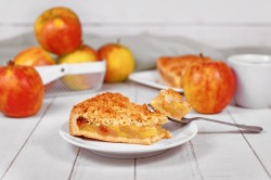 Ein Stück Apfelkuchen mit Zimtstreuseln auf einem Teller mit einer Kuchengabel, im Hintergrund Äpfel.
