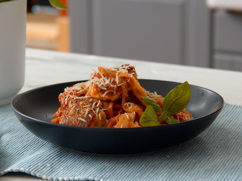 Ein schwarzer tiefer Teller mit Auberginen-Pasta auf einem Platzdeckchen auf einer Küchentheke.