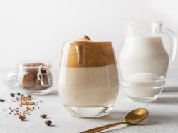 Ein Glas Dalgona Pumpkin Spice Latte, daneben ein goldener Löffel, eine Kanne Milch und eine Schale Kaffee.