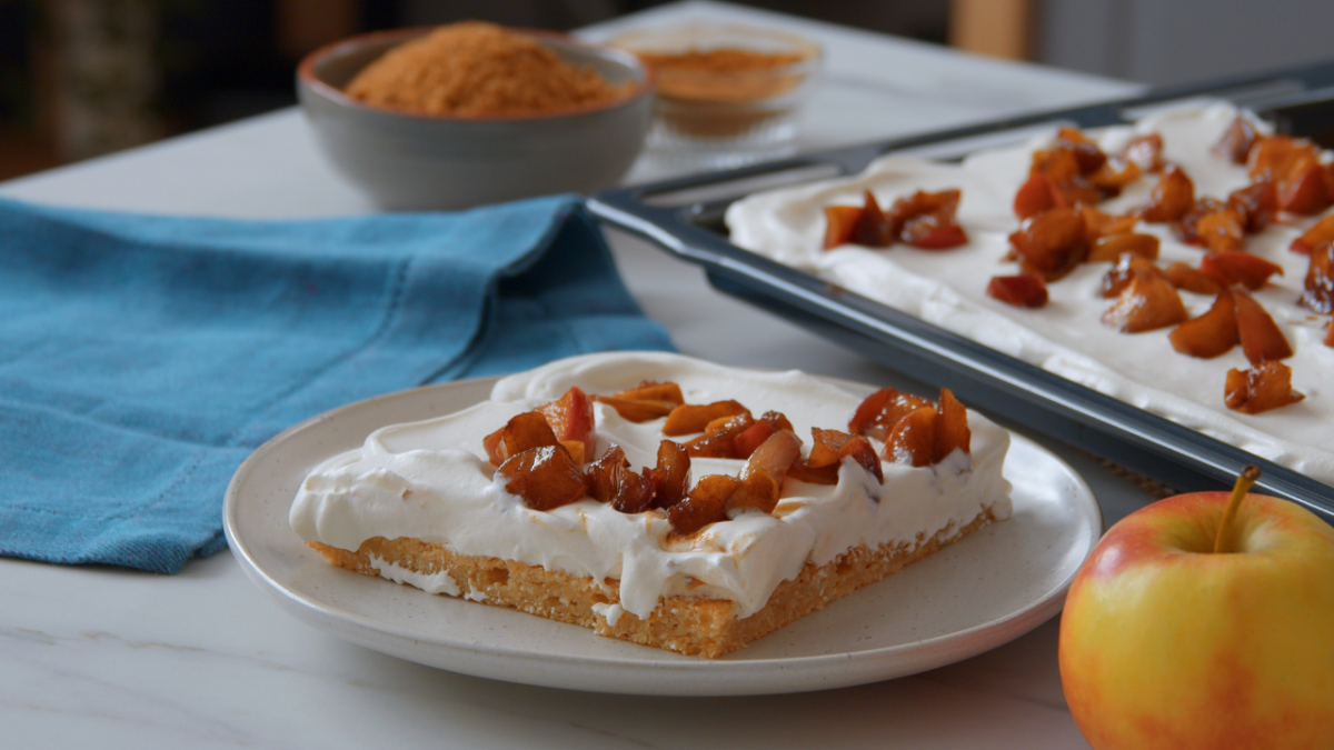 Ein Stück Apfelkuchen neben dem Blech mit Kuchen und einer Schüssel Zimt im Hintergrund.