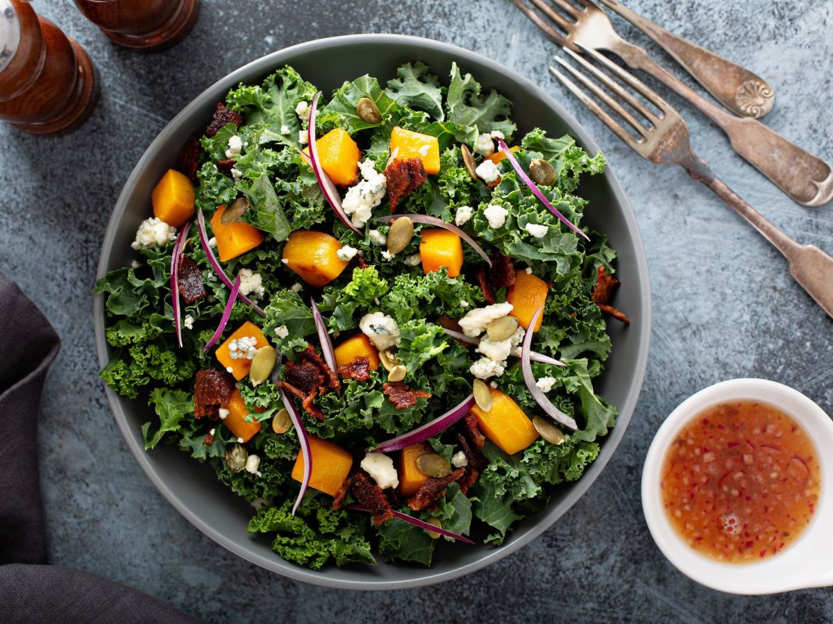 Bunter Grünkohlsalat mit geröstetem Kürbis, serviert in einer großen Schüssel. Ein Schälchen mit Dressing steht daneben. Im Hintergrund liegt Besteck