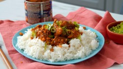 Eine Portion Hähnchencurry mit Reis auf Teller, daneben eine Schüssel mit geschnittenem Frühlingslauch und eine Dose mit Knuspererdnüssen.