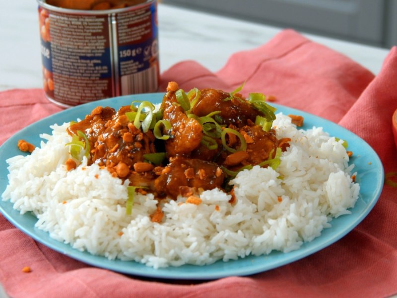 Eine Portion Hähnchencurry mit Reis auf Teller, daneben eine Schüssel mit geschnittenem Frühlingslauch und eine Dose mit Knuspererdnüssen.