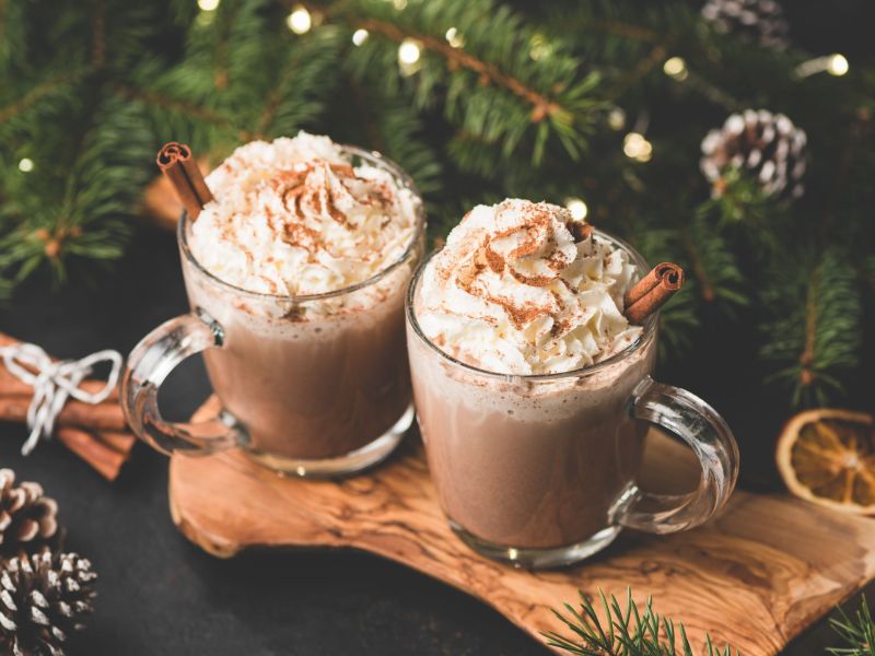 Zwei Becher mit heißer Schokolade mit Earl Grey stehen auf einem Holzbrett. Weihnachtliche Deko liegt davor und im Hintergrund.