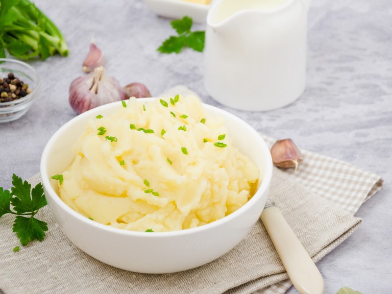Eine weiße Schüssel Knoblauch-Kartoffelpüree, daneben ein Löffel, Knoblauchzehen und Petersilie sowie ein Milchkännchen.