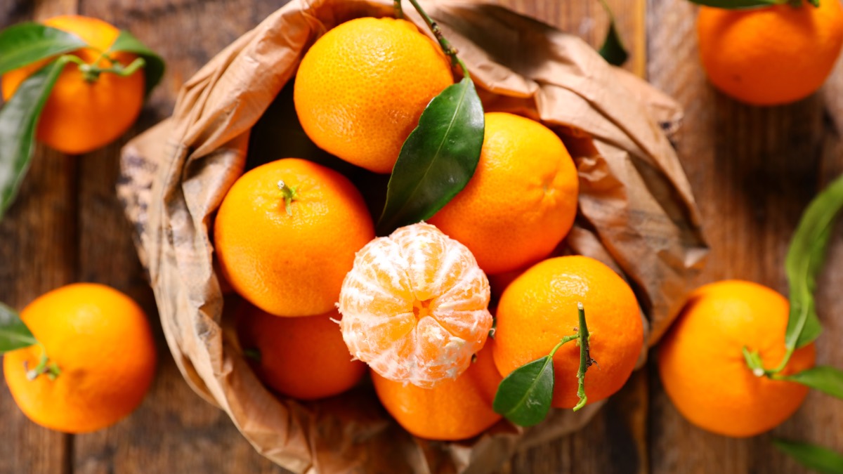 Eine Papiertüte voller Mandarinen. Eine abgepellte Mandarine thront obendrauf und mehrere Früchte liegen daneben.,