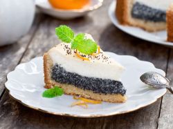 Ein Stück Mohn-Käse-Kuchen liegt auf einem weißen Teller mit Minzeblättchen, ein Teelöffel daneben. Ein weiteres Stück Kuchen steht im Hintergrund.