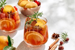 zwei Gläser weihnachtlicher Zitrus-Cocktail mit Zitrusscheiben, Zimtstangen, Sternanis und Rosmarin, daneben liegen Sternanis und Zimtstangen, im Hintergrund eine Schale mit Obstscheiben.