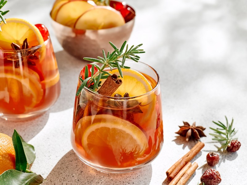 zwei Gläser weihnachtlicher Zitrus-Cocktail mit Zitrusscheiben, Zimtstangen, Sternanis und Rosmarin, daneben liegen Sternanis und Zimtstangen, im Hintergrund eine Schale mit Obstscheiben.