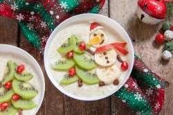 Zwei Schüsseln Weihnachtsmilchreis dekoriert mit Schneemännern und Weihnachtsbäumen aus Bananen und Kiwi, drumherum weihnachtliche Deko, Draufsicht.