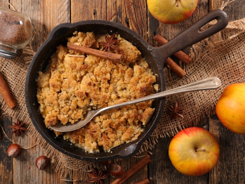 Eine Pfanne mit Apfel-Stollen-Crumble in der Draufsicht. Daneben liegen Äpfel und Zimtstange. Im Crumble liegt ein Servierlöffel.