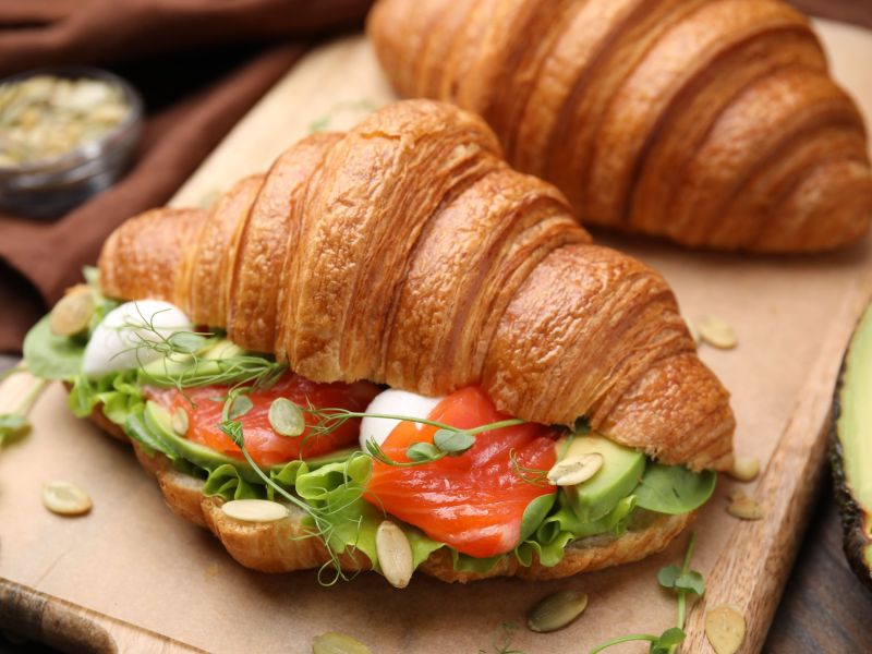 Zwei Croissants liegen auf einem Holzbrett. Das vordere Croissant ist aufgeschnitten. Gefüllt ist das Avocado-Lachs-Croissant mit Avocado, Räucherlachs und Mozzarella. Einige Salatblätter, Sprossen und Kürbiskerne dienen als Dekoration.