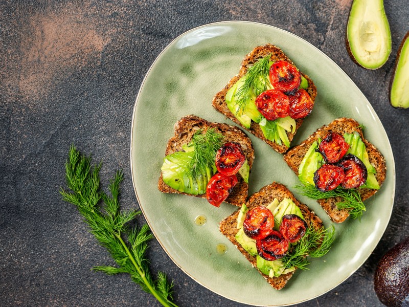 3 Avocado-Brote mit Ofentomaten und Kräutern belegt auf einem Teller in der Draufsicht, drumherum ganze und halbierte Avocados.