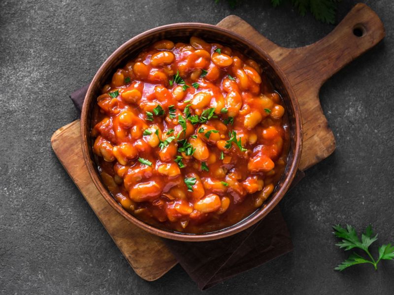 Baked Beans, serviert in einer gusseisernen Pfanne. Frische Kräuter als Deko. Die Pfanne steht auf einem Holzbrett.