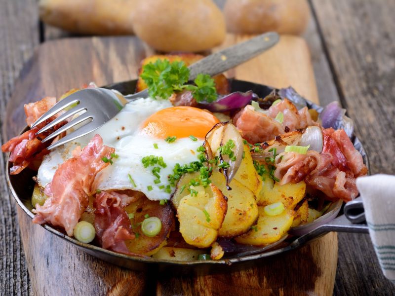 Deftiges Bauernfrühstück mit Kartoffeln, Speck und Spiegelei, serviert in einer gusseisernen Pfanne. Daneben liegt eine Gabel.