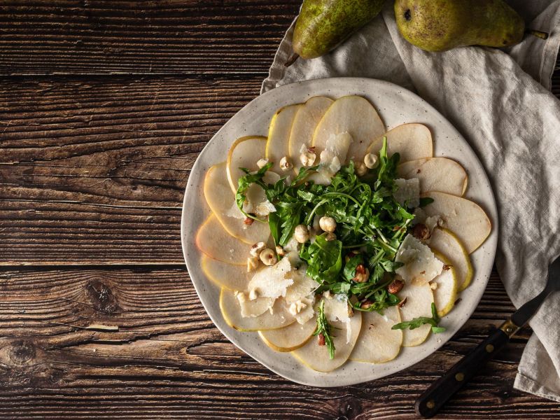 Birnen-Carpaccio mit Gorgonzola und Walnüssen arrangiert auf einem Teller. Eine helle Tischdecke liegt daneben.