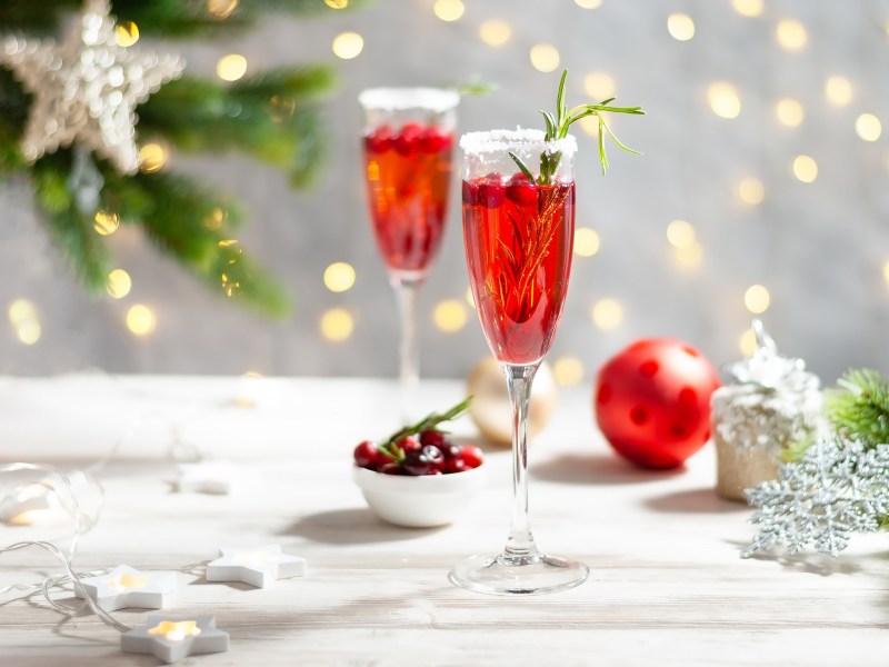 2 Gläser Cranberry Mimosas mit Cranberrys und Rosmarin garniert, im Hintergrund weihnachtliche Deko.