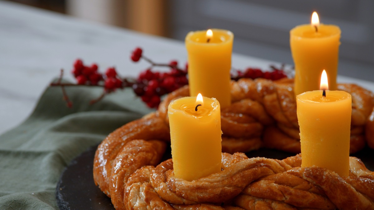 Ein essbarer Adventskranz aus Brezelteig mit vier Kerzen aus Butter. Im Hintergrund eine grüne Serviette und rote Pflanzen, passend zu Weihnachten.