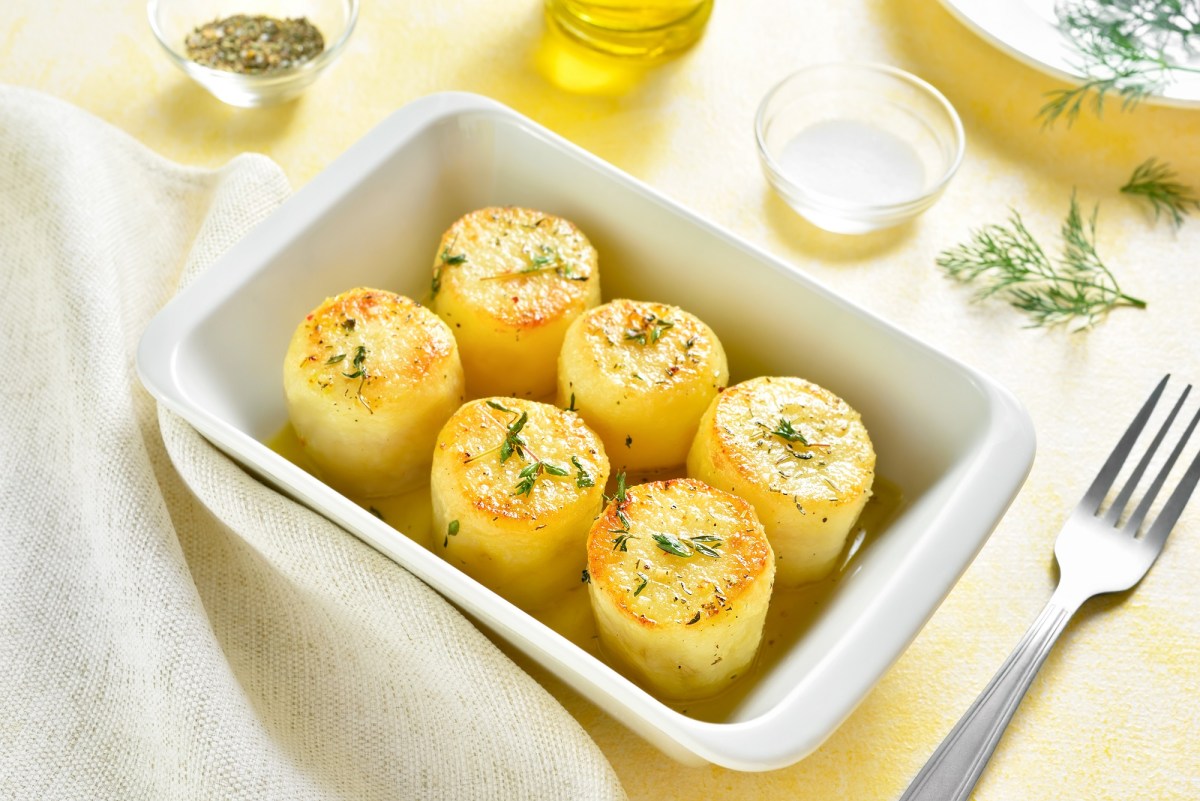 Eine weiße rechteckige Auflaufform mit Fondant-Kartoffeln, bestreut mit Rosmarin, daneben eine Gabel und ein paar Zutaten wie Brühe und flüssige Butter.