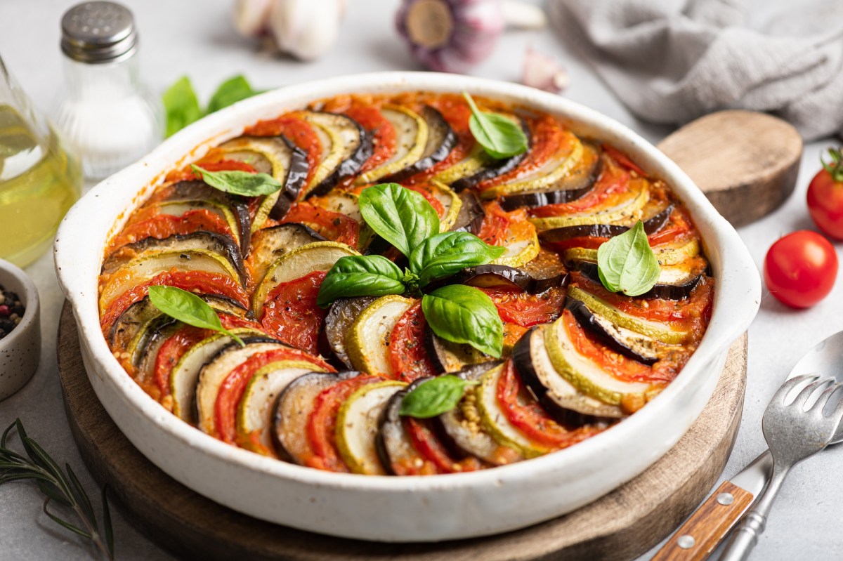 französischer Gemüseauflauf Tian de Légumes in einer runden Auflaufform auf einem Holzbrett, drumherum viele Zutaten.