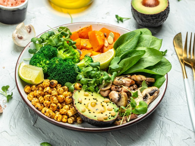 Eine herzhafte Frühstücksbowl mit Spinat, Avocado, Pilzen, Kichererbsen, Brokkoli und Süßkartoffel, daneben Gabel und Messer, außerdem Zutaten.