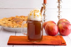 Ein Glas Hot Apple Pie garniert mit geschlagener Sahne, Karamellsoße und einer Zimtstange, auf einem Brett, dahinter ganze Äpfel und ein Apfel-Pie.