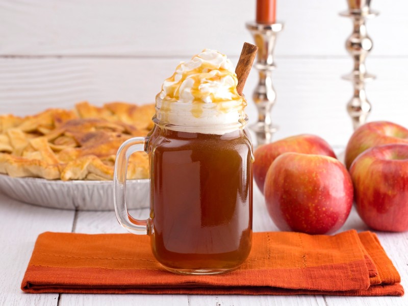 Ein Glas Hot Apple Pie garniert mit geschlagener Sahne, Karamellsoße und einer Zimtstange, auf einem Brett, dahinter ganze Äpfel und ein Apfel-Pie.
