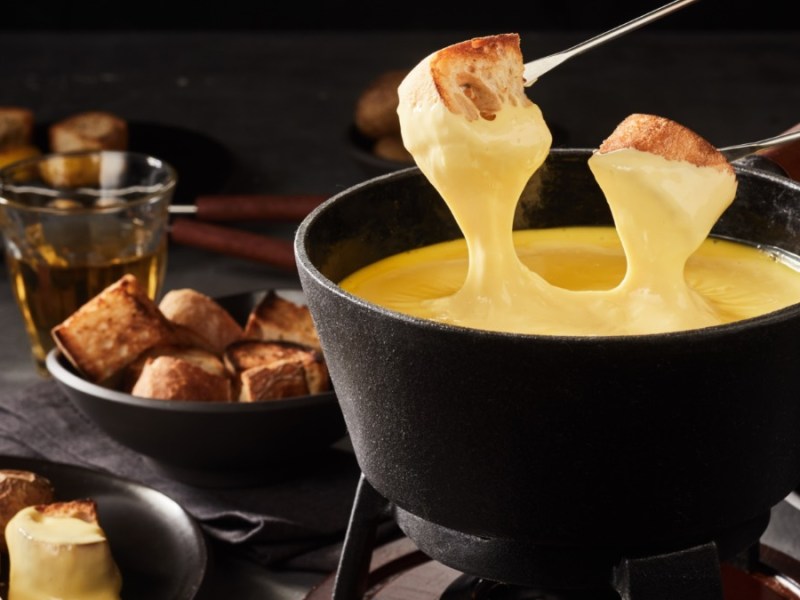 Ein Käsefondue, in das zwei Brotstücke getaucht werden. Im Hintergrund stehen Schalen und Teller mit Brot und ein Glas mit Wein.