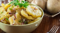 Eine Schüssel mit Kartoffelsalat, garniert mit Petersilie. Daneben liegt eine Gabel und rohe Kartoffeln im Hintergrund.