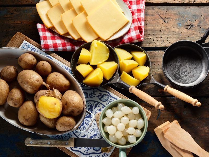Raclette-Zutaten wie Käse, Kartoffeln und Silberzwiebeln sind zu sehen. Daneben liegen ein scharfest Messer, einige Holzschieber. Aus übrig gebliebenen Zutaten kann man wunderbar Raclette-Reste verarbeiten.