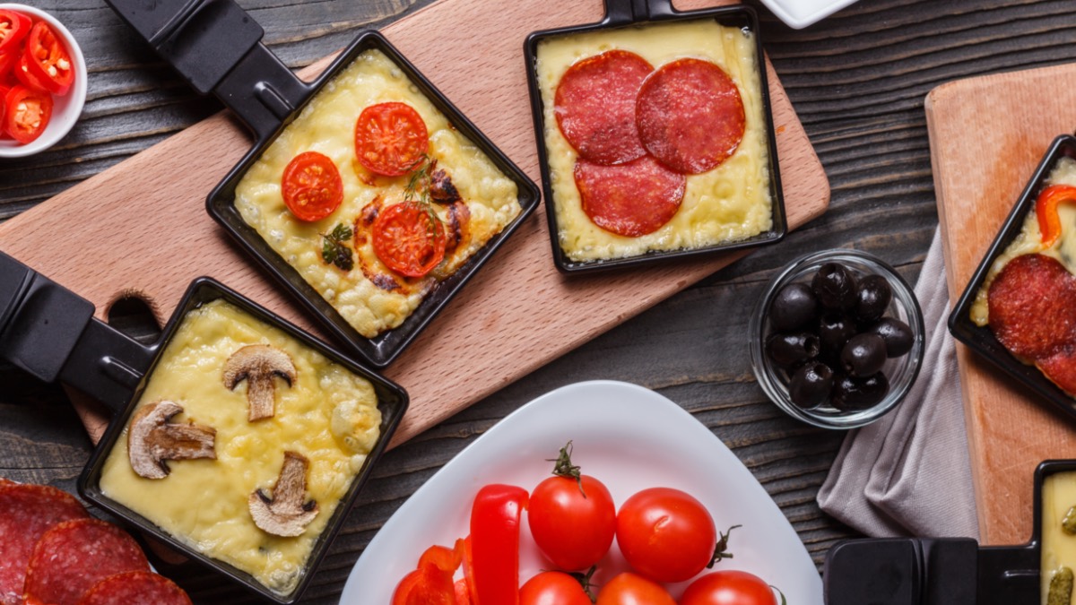 Drei Raclette-Pfännchen, die unterschiedlich gefüllt sind, in der Draufsicht. Drumherum liegen Tomaten und weitere Zutaten.