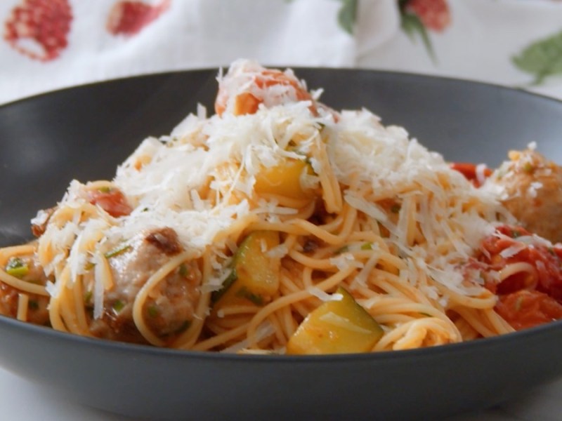 Ein schqarzer Teller mit einer Pasta-Pfanne mit Fleischbällchen, Gemüse und geriebenem Parmesan.