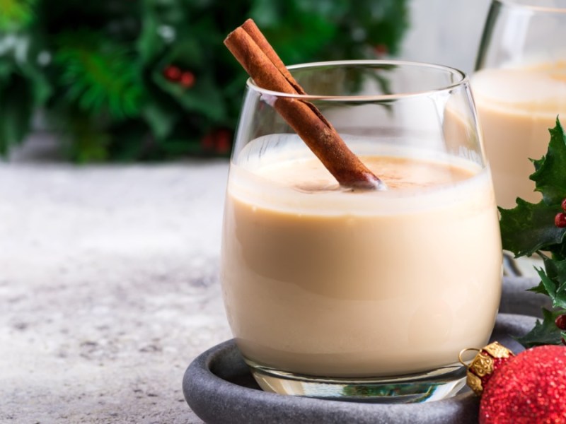 Ein Glas mit Spekulatius-Likör, in dem eine Zimtstange steckt. Im Hintergrund steht ein weiteres Glas, einige Zweige Stechpalme und eine Christbaumkugel.