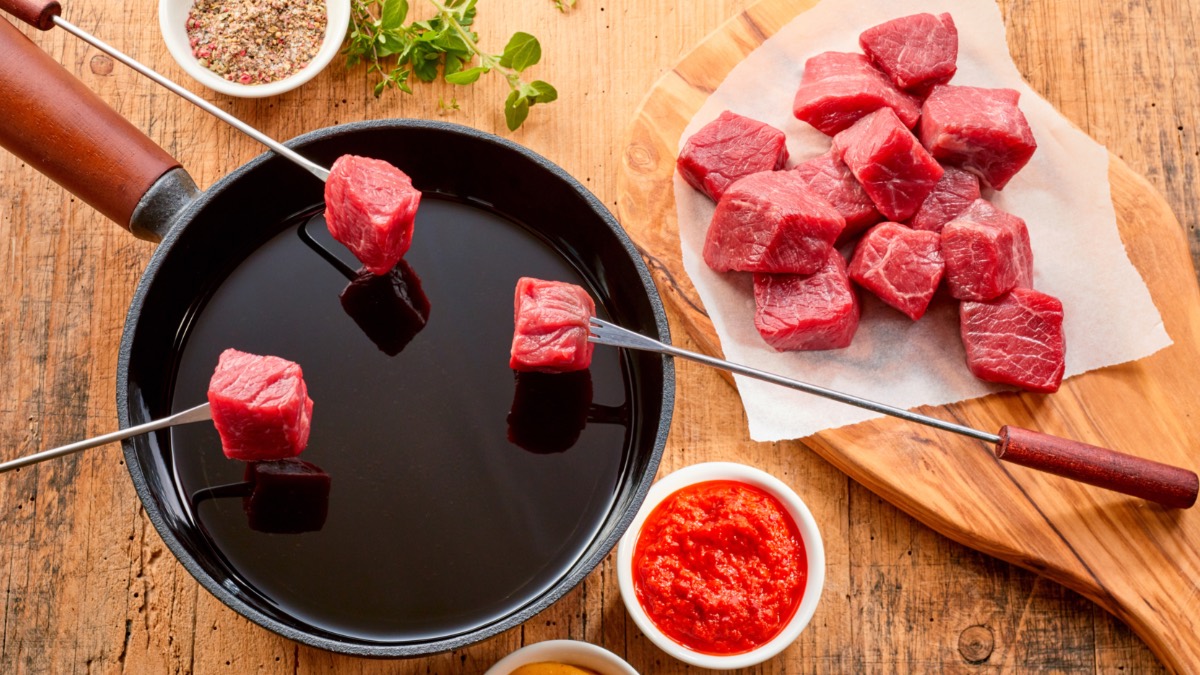 Ein Fondue, übder das 3 Fonduegabeln mit Fleisch gehalten werden, in der Draufsicht. Daneben Schälchen mit Soßen und ein Teller voller Fleisch.