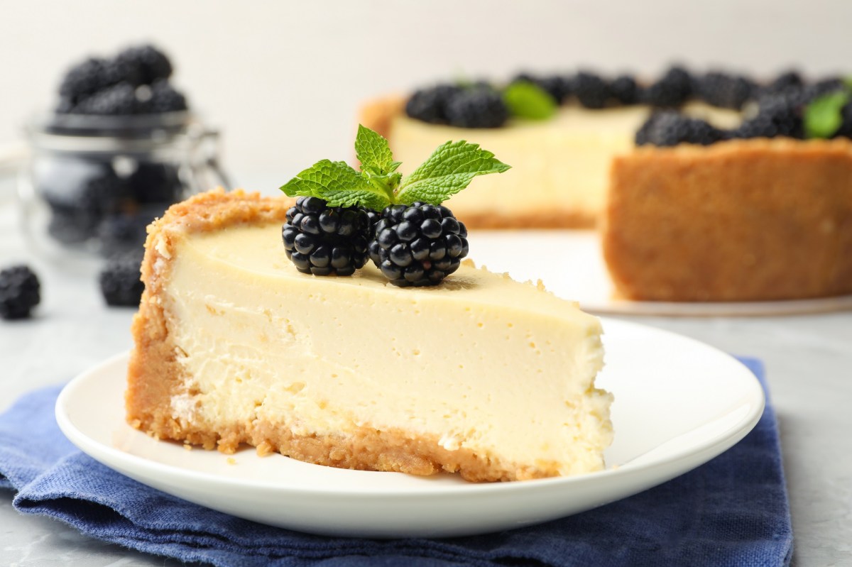 Ein Stück veganer Käsekuchen auf einem Teller, garniert mit Minze und Brombeeren, dahinter unscharf der angeschnittene Kuchen.