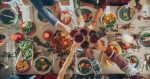Ein weihnachtlich gedeckter Esstisch mit vielen gefüllten Schalen mit Essen, über dem eine Gruppe von Menschen mit roten Weihnachtscocktails im Glas anstößt. Draufsicht.