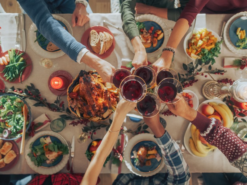 Ein weihnachtlich gedeckter Esstisch mit vielen gefüllten Schalen mit Essen, über dem eine Gruppe von Menschen mit roten Weihnachtscocktails im Glas anstößt. Draufsicht.
