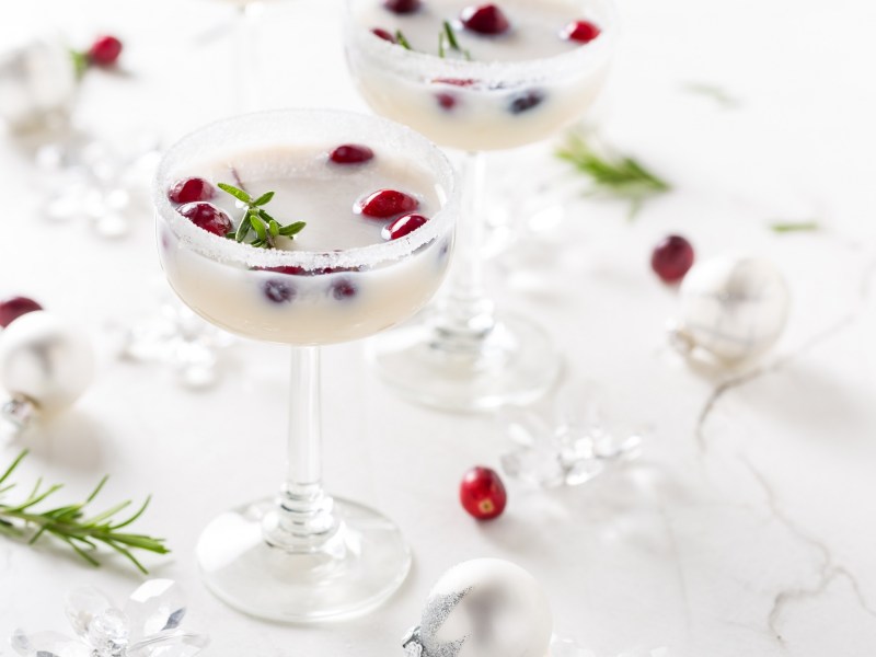 2 Gläser White Christmas Margarita mit Kokosrand und frischen Cranberrys garniert, daneben Weihnachtsdeko und Cranberrys.