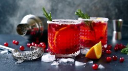 2 Gläser Winter-Margarita mit Rosmarinzwigen. Im Hintergrund ein Cocktailshaker, Eiswürfel, Cranberrys, Orangen und Rosmarinzweige.