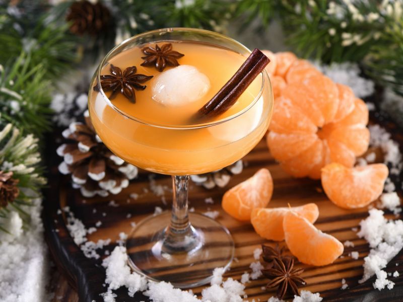 Winter-Old Fashioned, serviert in einem bauchigen Glas. Zimtstange und Sternanis als Deko. Im Hintergrund liegen Mandarinenstücke und eine Mandarine als Deko sowie einge Tannenzapfen.