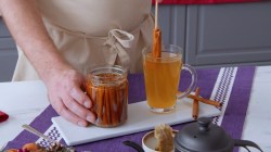 Ein Honig-Glas voller Zimtstangen auf der linken Seite und daneben ein Glas Tee, in den eine Honig-Zimtstange gegeben wird.