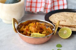 Eine Schüssel Aloo Gobi mit Blumenkohl und Kartoffeln, garniert mit einer Zitronenscheibe, daneben ein Teller mit Fladenbrot.