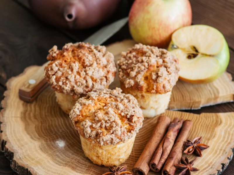 3 Apfel-Streusel-Muffins auf einer runden Holzplatte, daneben Äpfel und Zimtstangen.