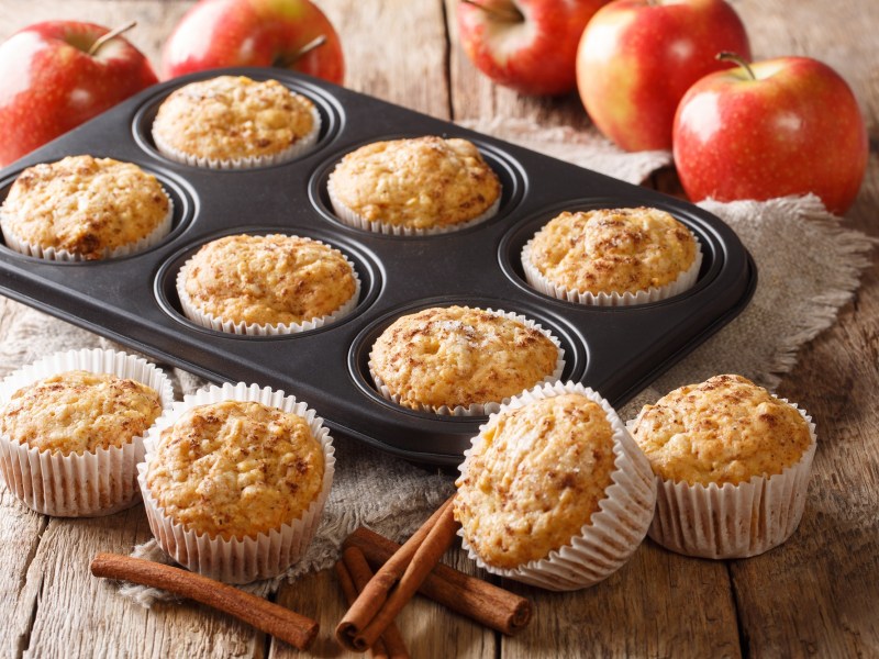 Ein Muffinblech mit Apfelmus-Muffins, drumherum weitere Muffins und frische Äpfel sowie Zimtstangen.