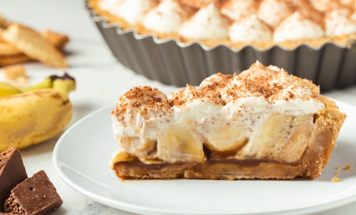Ein Stück Banoffee Pie auf einem Teller. Daneben liegen Banane und der Rest der Pie ist im Hintergrund zu sehen.