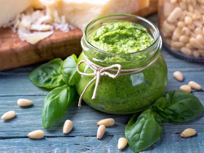 Ein Glas Basilikumpesto verziert mit einer Schleife, darum herum Basilikumblätter und Pinienkerne.