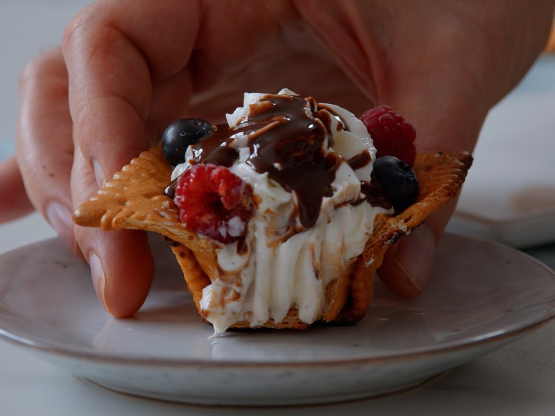 Ein angebissener Butterkeks-Muffin mit Vanillesahne, Beeren und Schokosoße, der auf einen Teller gesetzt wird.