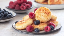 Ein Stapel Hüttenkäse-Pancakes mit frischen Himbeeren und Blaubeeren auf einem Teller. Im Hintergrund sind Schalen mit den Beeren und weitere Pancakes zu sehen.