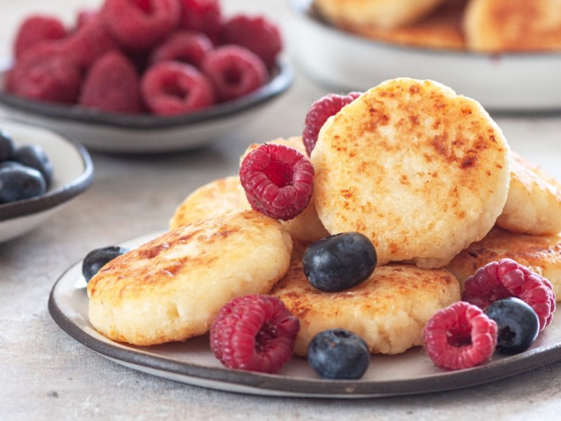 Ein Stapel Hüttenkäse-Pancakes mit frischen Himbeeren und Blaubeeren auf einem Teller. Im Hintergrund sind Schalen mit den Beeren und weitere Pancakes zu sehen.